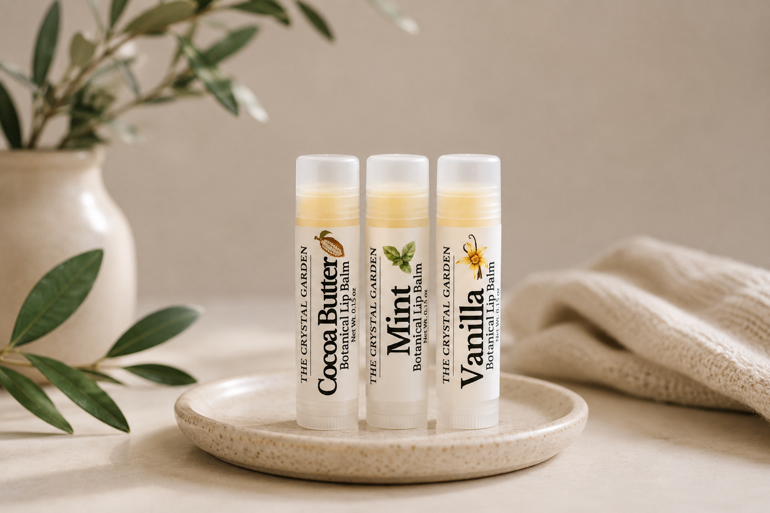Three lip balm tubes from The Crystal Garden stand on a small round tray. The tubes are labeled Cocoa Butter, Mint, and Vanilla, with white caps and clear tops showing pale yellow balm inside.