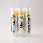 Three tubes of botanical lip balm from The Crystal Garden lined up on a light gray background. The tubes are labeled Citrus Mint, Mint, and Sweet Mint, with corresponding citrus and mint illustrations on the white labels.