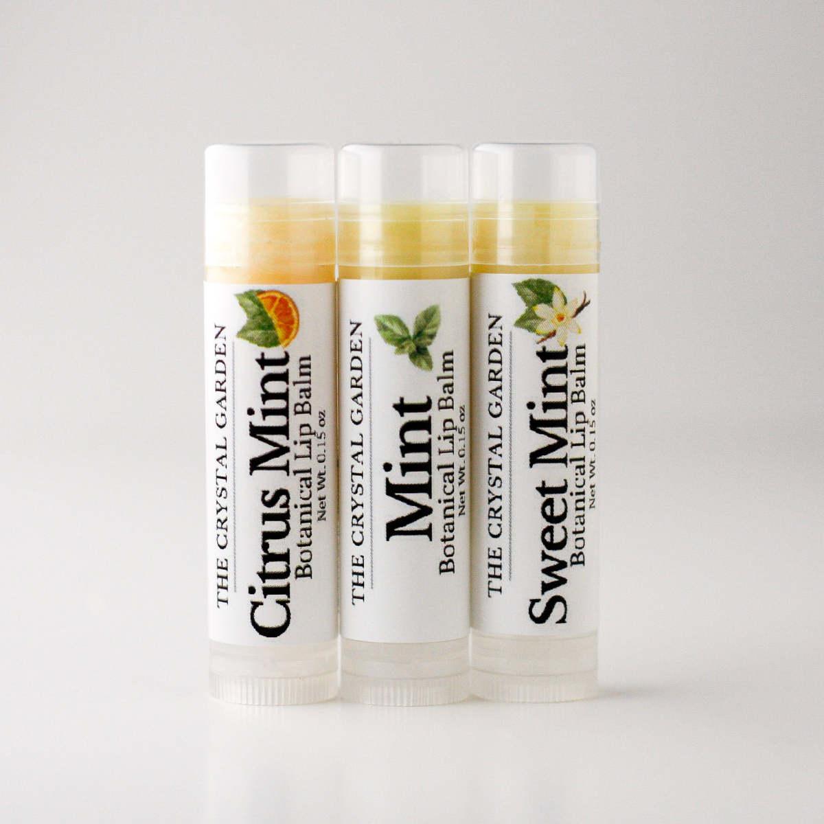 Three tubes of botanical lip balm from The Crystal Garden lined up on a light gray background. The tubes are labeled Citrus Mint, Mint, and Sweet Mint, with corresponding citrus and mint illustrations on the white labels.