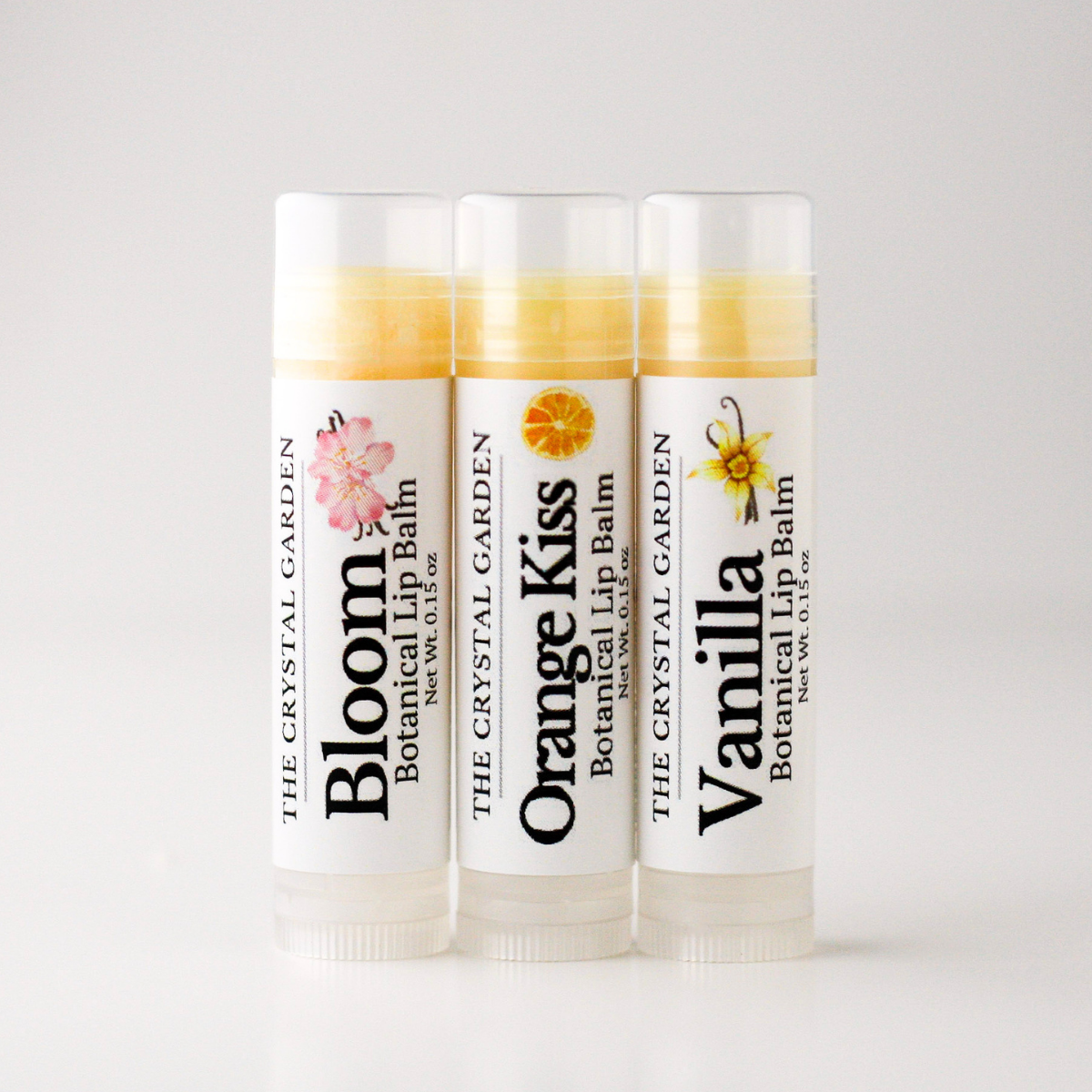 Three tubes of botanical lip balm from The Crystal Garden lined up on a light gray background. The tubes are labeled Bloom, Orange Kiss, and Vanilla, with white caps and white labels featuring colorful botanical illustrations.