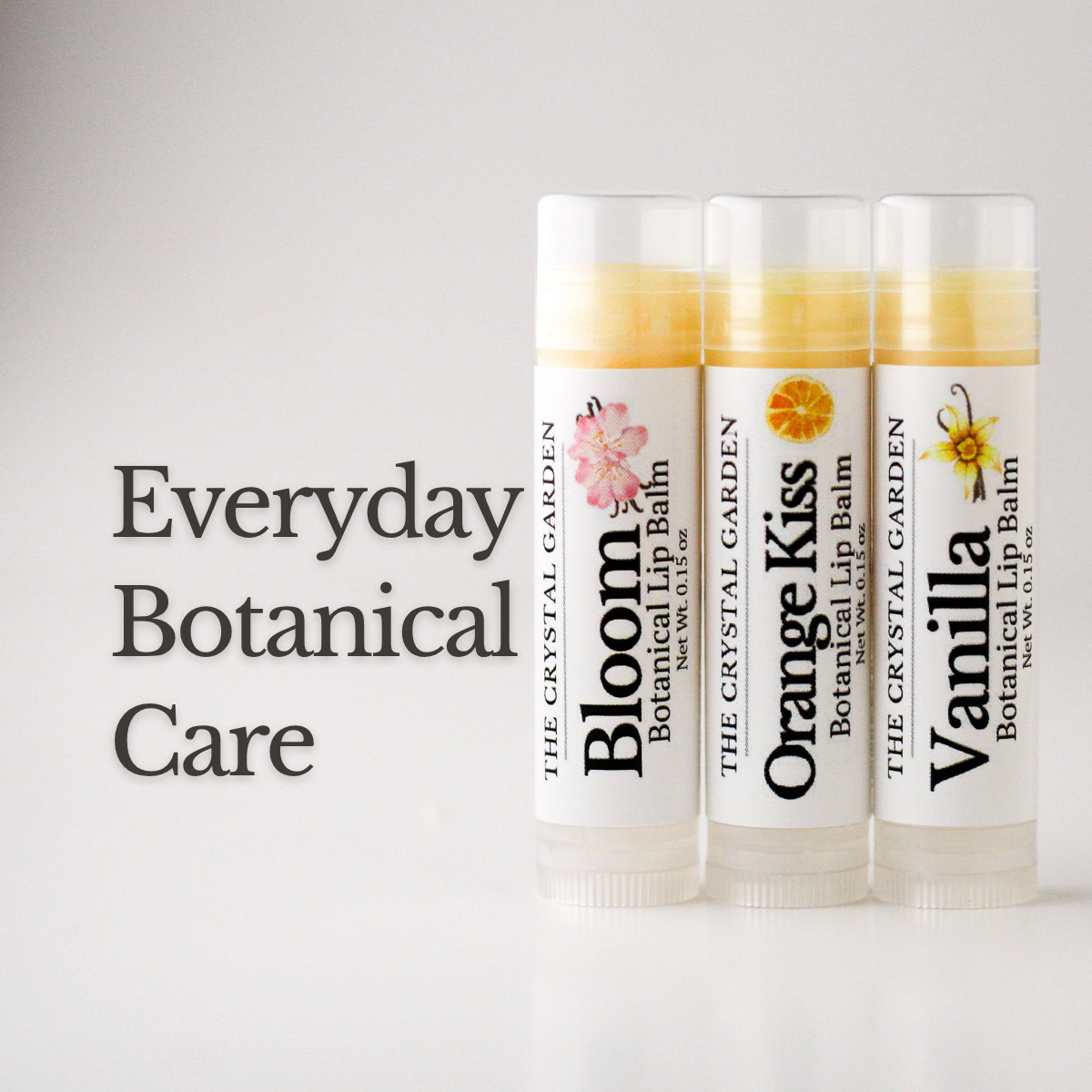 Three tubes of botanical lip balm from The Crystal Garden are lined up on a white background next to the text 'Everyday Botanical Care'. The tubes are labeled Bloom, Orange Kiss, and Vanilla, with white caps and clear lower sections showing the yellow balm inside.