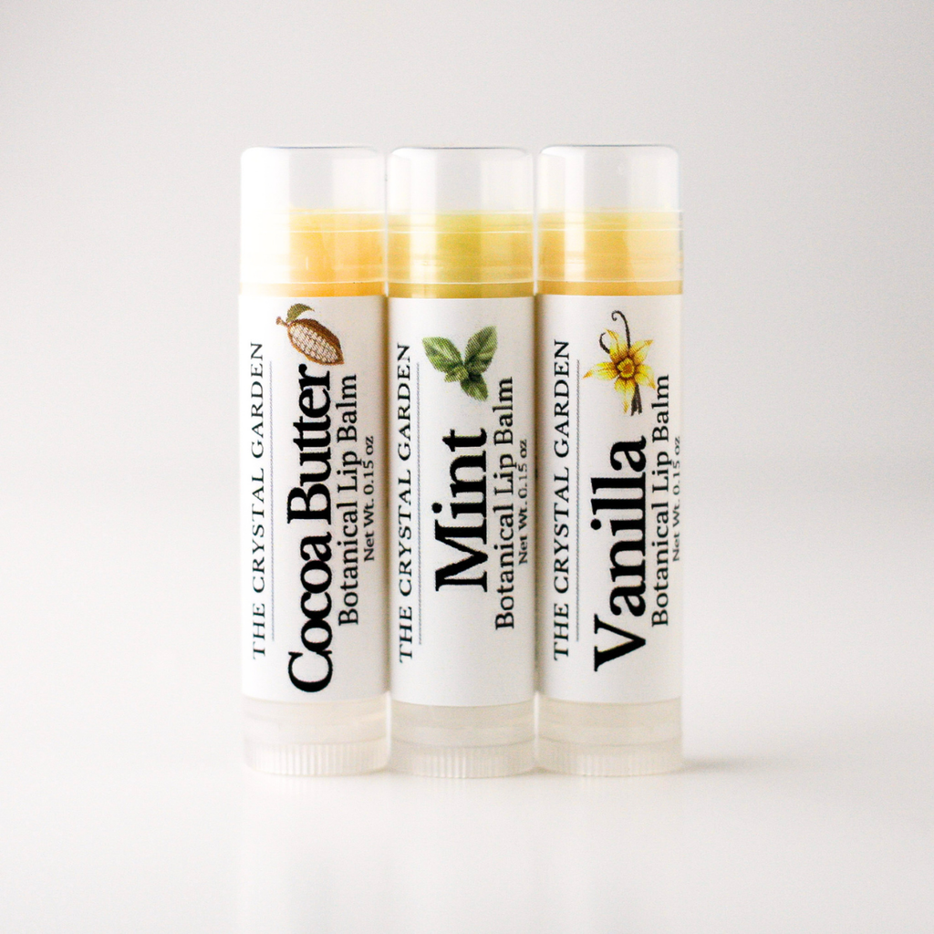 Three tubes of botanical lip balm from The Crystal Garden lined up on a white background, labeled Cocoa Butter, Mint, and Vanilla.