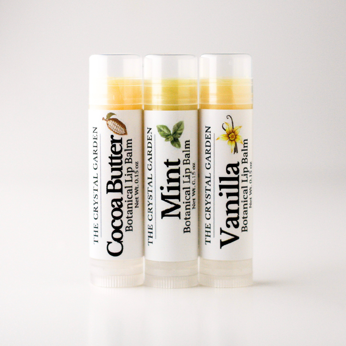 Three tubes of botanical lip balm from The Crystal Garden lined up on a white background, labeled Cocoa Butter, Mint, and Vanilla.