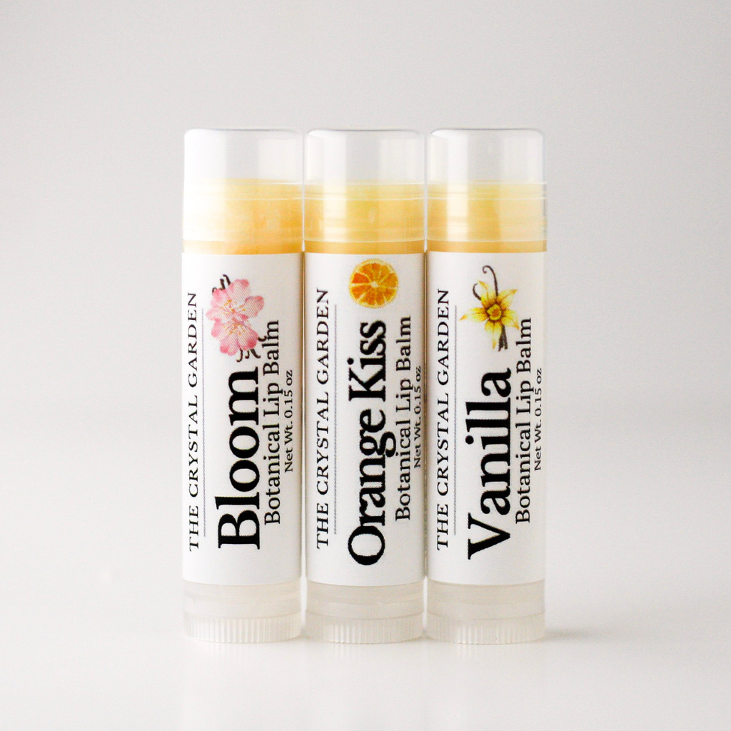 Three tubes of botanical lip balm from The Crystal Garden lined up on a light gray background. The tubes are labeled Bloom, Orange Kiss, and Vanilla, with white caps and white labels featuring colorful botanical illustrations.