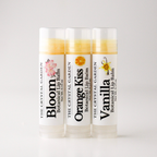 Three tubes of botanical lip balm from The Crystal Garden lined up on a light gray background. The tubes are labeled Bloom, Orange Kiss, and Vanilla, with white caps and white labels featuring colorful botanical illustrations.