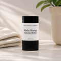 A black and white stick of 'The Crystal Garden' Baby Bump Botanical Butter is centered on a light marble surface, flanked by a folded cream towel on the left and a potted plant on the right.