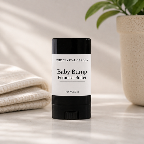 A black and white stick of 'The Crystal Garden' Baby Bump Botanical Butter is centered on a light marble surface, flanked by a folded cream towel on the left and a potted plant on the right.