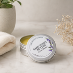An open round silver tin of Baby Tushy Butter Cocoa Butter by The Crystal Garden sits on a light marble surface next to a folded cream towel, with a potted plant and dried baby's breath in the background.