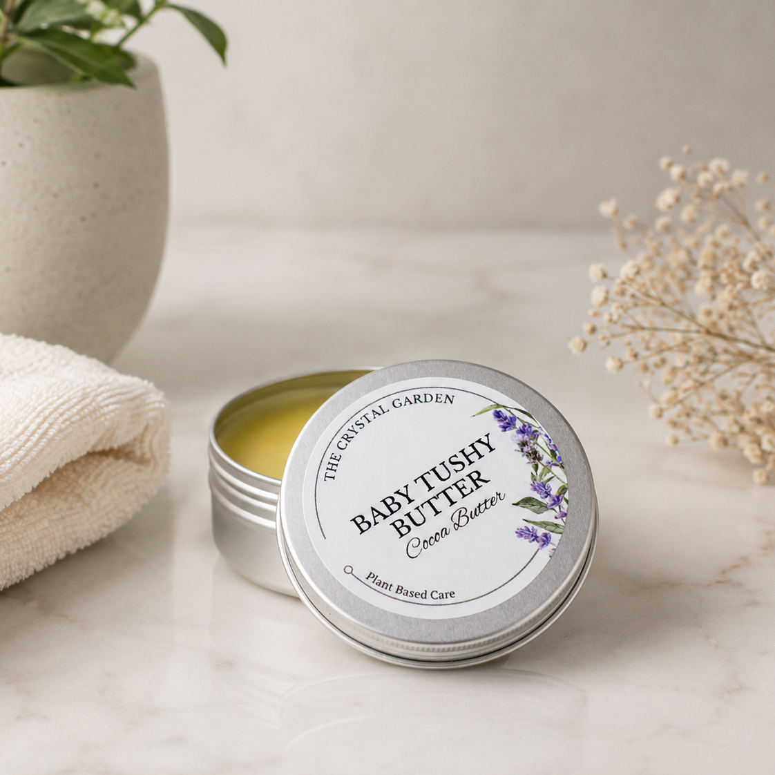 An open round silver tin of Baby Tushy Butter Cocoa Butter by The Crystal Garden sits on a light marble surface next to a folded cream towel, with a potted plant and dried baby's breath in the background.