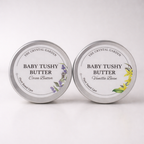 Two round silver metal tins of Baby Tushy Butter by The Crystal Garden, shown side by side on a white background. The left tin is labeled Cocoa Butter with purple lavender illustrations, and the right tin is labeled Vanilla Bean with yellow flower illustrations.