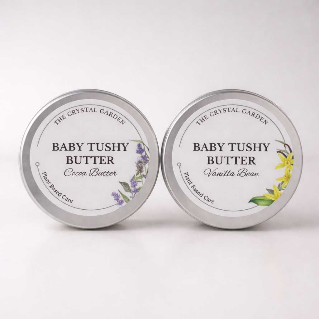 Two round silver metal tins of Baby Tushy Butter by The Crystal Garden, shown side by side on a white background. The left tin is labeled Cocoa Butter with purple lavender illustrations, and the right tin is labeled Vanilla Bean with yellow flower illustrations.