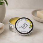 An open silver tin of Baby Tushy Butter Vanilla Bean balm sits on a light marble surface, with its lid leaning beside it. The round lid has a black border and a white label with yellow flower illustrations.