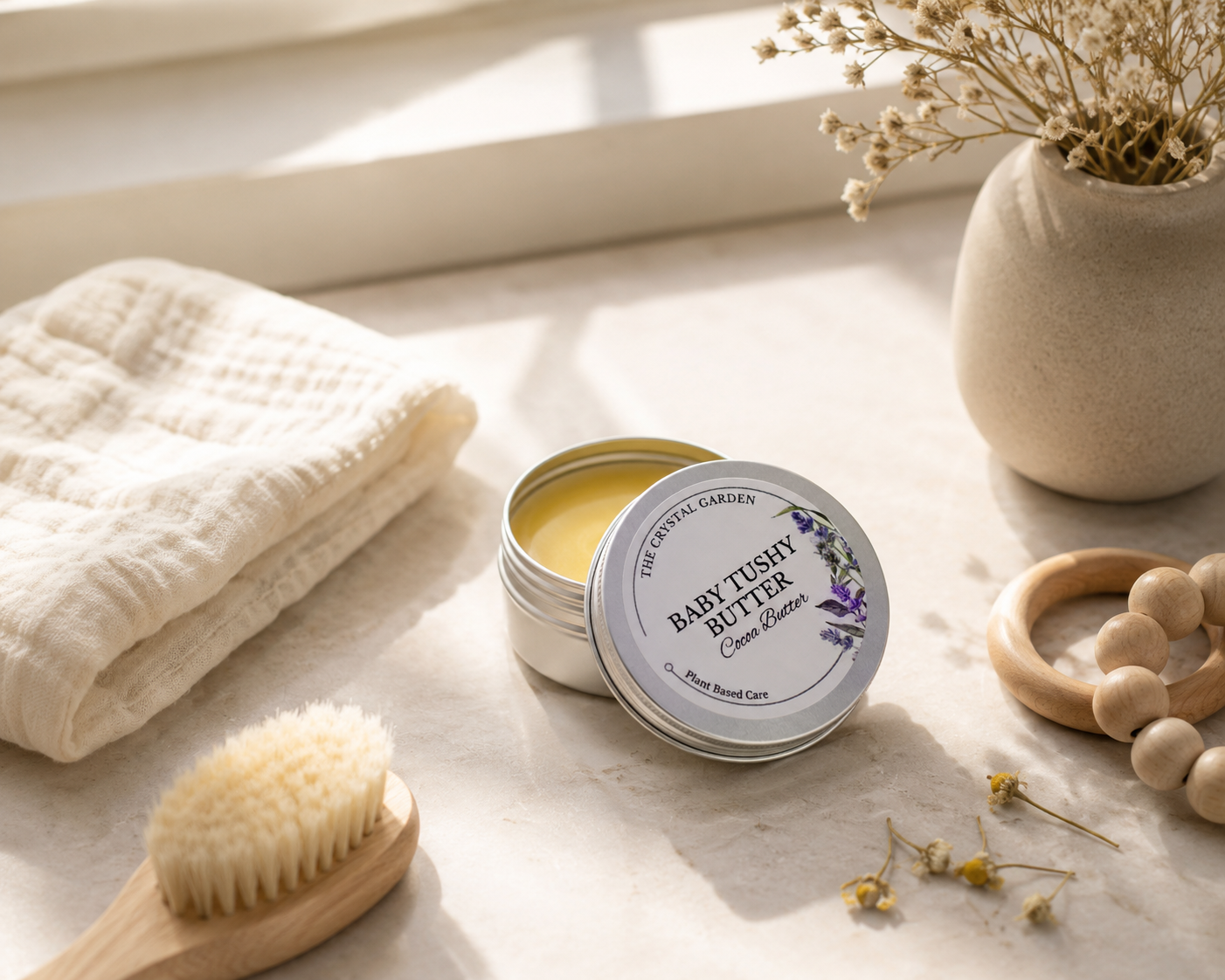 Open round tin of Baby Tushy Butter body butter on a light stone surface, surrounded by a folded cream towel, wooden brush, wooden beads, and dried flowers in a vase.