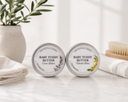 Two round silver tins of The Crystal Garden Baby Tushy Butter sit on a white marble surface with folded white towels, a plant in a vase, and a wooden brush in the background. The left tin is labeled Cream Butter with lavender illustrations, and the right tin is labeled Vanilla Rose with yellow floral illustrations.