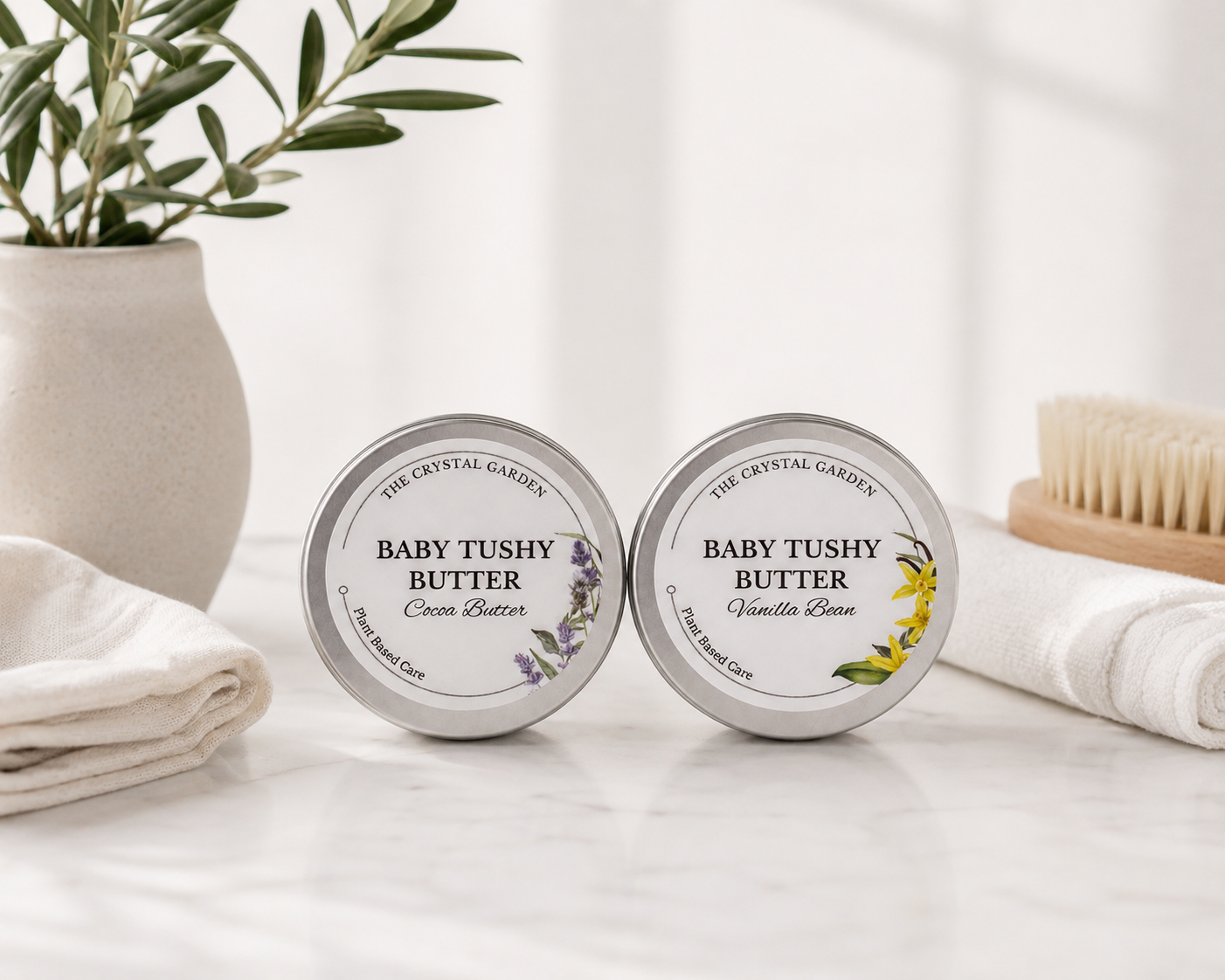 Two round silver tins of The Crystal Garden Baby Tushy Butter sit on a white marble surface with folded white towels, a plant in a vase, and a wooden brush in the background. The left tin is labeled Cream Butter with lavender illustrations, and the right tin is labeled Vanilla Rose with yellow floral illustrations.