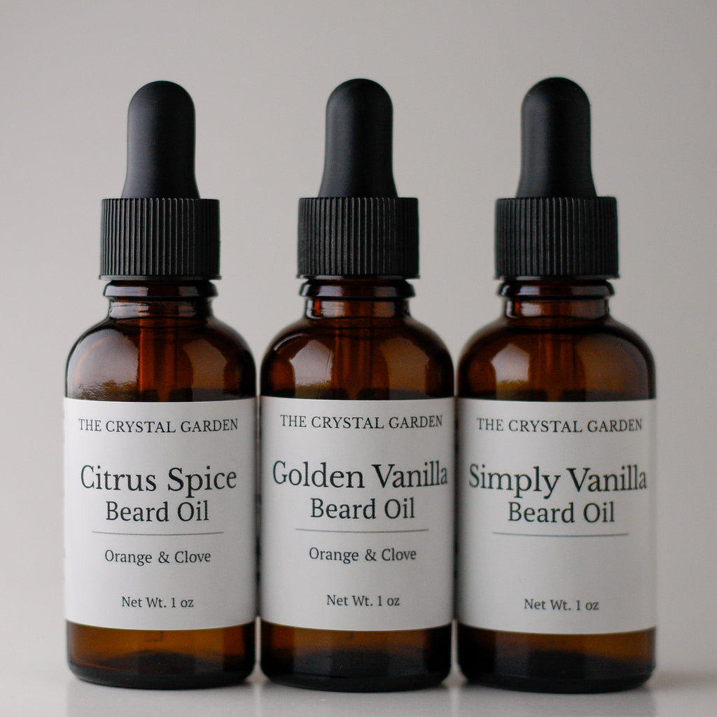 Three amber glass dropper bottles of beard oil from The Crystal Garden, arranged side by side on a light gray background. The bottles are labeled Citrus Spice Beard Oil, Golden Vanilla Beard Oil, and Simply Vanilla Beard Oil.