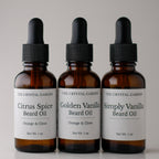 Three amber glass dropper bottles of beard oil from The Crystal Garden, arranged side by side on a light gray background. The bottles are labeled Citrus Spice Beard Oil, Golden Vanilla Beard Oil, and Simply Vanilla Beard Oil.