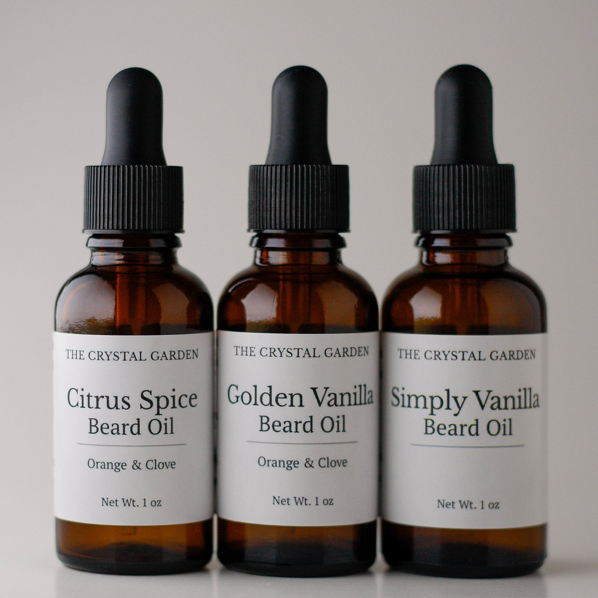 Three amber glass dropper bottles of beard oil from The Crystal Garden, arranged side by side on a light gray background. The bottles are labeled Citrus Spice Beard Oil, Golden Vanilla Beard Oil, and Simply Vanilla Beard Oil.