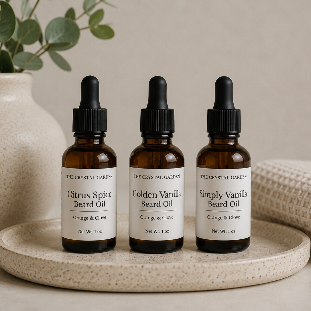 Three amber glass dropper bottles of beard oil from The Crystal Garden are arranged on a round beige tray. The bottles are labeled Citrus Spice Beard Oil, Golden Vanilla Beard Oil, and Simply Vanilla Beard Oil, with orange and clove notes.