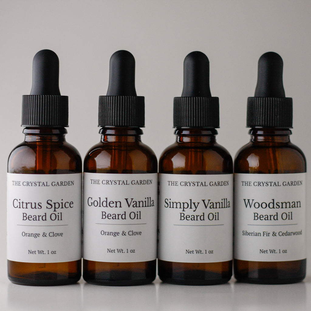 Four amber glass dropper bottles of beard oil from The Crystal Garden lined up on a light gray background. The labels read: Citrus Spice Beard Oil, Golden Vanilla Beard Oil, Simply Vanilla Beard Oil, and Woodsman Beard Oil.