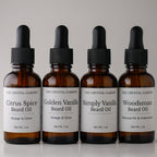 Four amber glass dropper bottles of beard oil from The Crystal Garden lined up on a light gray background. The labels read: Citrus Spice Beard Oil, Golden Vanilla Beard Oil, Simply Vanilla Beard Oil, and Woodsman Beard Oil.