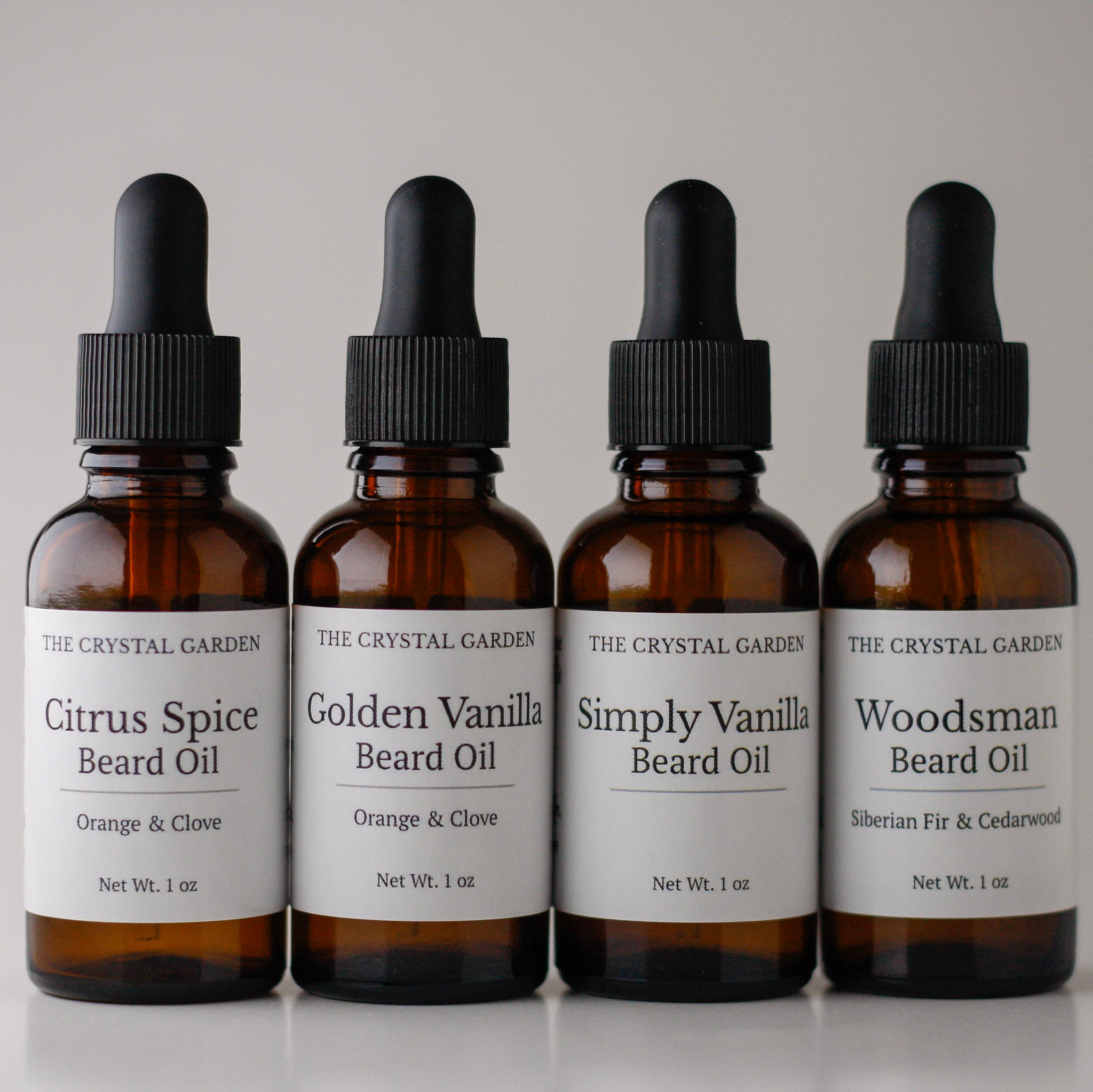 Four amber glass dropper bottles of beard oil from The Crystal Garden lined up on a light gray background. The labels read: Citrus Spice Beard Oil, Golden Vanilla Beard Oil, Simply Vanilla Beard Oil, and Woodsman Beard Oil.