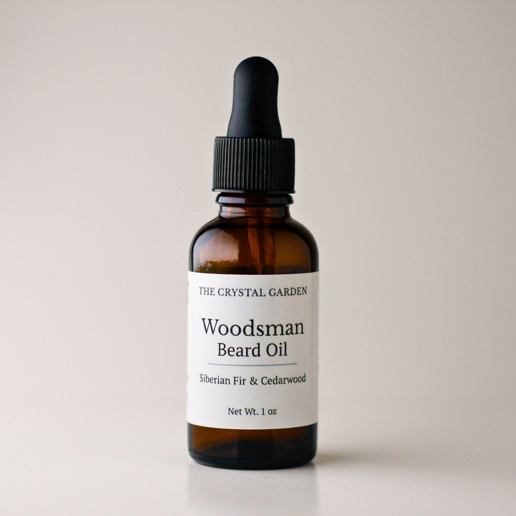 A 1 oz amber glass dropper bottle of The Crystal Garden Woodsman Beard Oil with a black cap and white label, set against a plain light beige background.