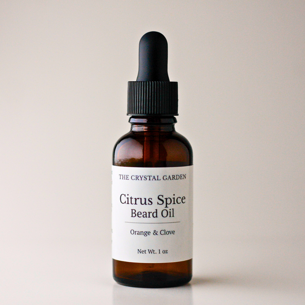 An amber glass dropper bottle of The Crystal Garden Citrus Spice Beard Oil with a black dropper cap and a white label, set against a solid light beige background.