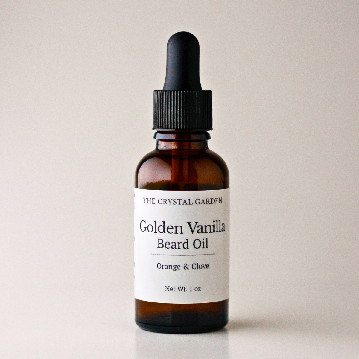 An amber glass dropper bottle of Golden Vanilla Beard Oil by The Crystal Garden, featuring a white label and a black rubber dropper cap, set against a plain light beige background.