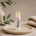 A tube of Bloom Botanical Balm by The Crystal Garden stands upright on a small round ceramic dish. The clear cap reveals a pale yellow balm inside, and the label is white with pink floral graphics and black text.