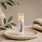 A tube of Bloom Botanical Balm by The Crystal Garden stands upright on a small round ceramic dish. The clear cap reveals a pale yellow balm inside, and the label is white with pink floral graphics and black text.
