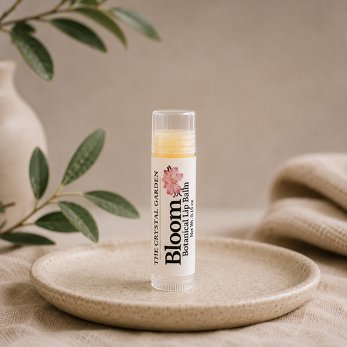 A tube of Bloom Botanical Balm by The Crystal Garden stands upright on a small round ceramic dish. The clear cap reveals a pale yellow balm inside, and the label is white with pink floral graphics and black text.