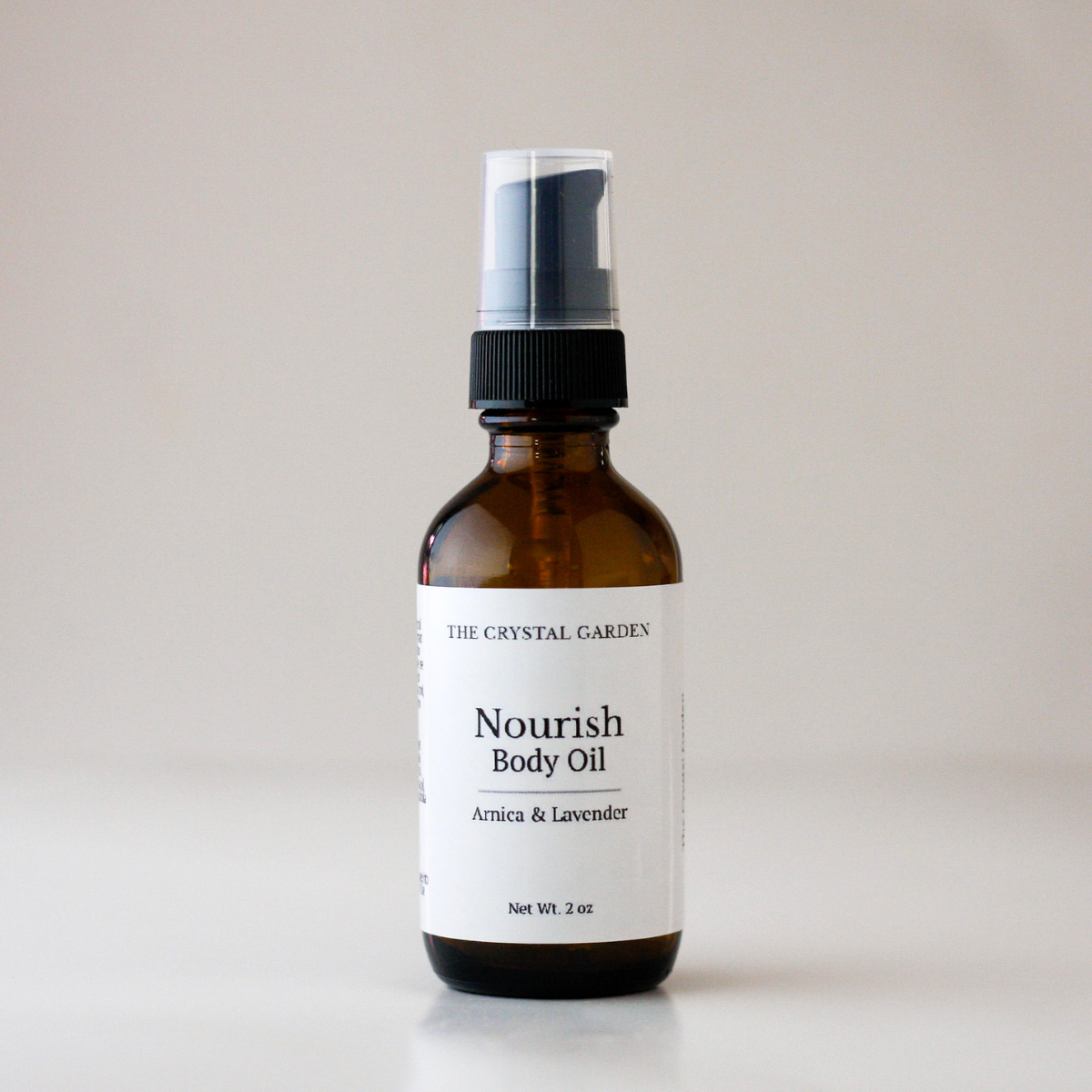 An amber glass spray bottle of Nourish Body Oil by The Crystal Gardex, featuring a white label and a black spray nozzle, set against a neutral light gray background.