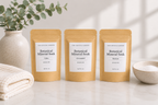 Three stand-up pouches of The Crystal Garden Botanical Mineral Soak arranged on a light marble surface with a folded cream towel, a ceramic vase with greenery, and a small bowl in the background.