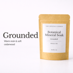 A single-use kraft paper pouch of The Crystal Garden Grounded Botanical Mineral Soak, shown next to text describing the scent as warm resin and soft cedarwood on a white background.