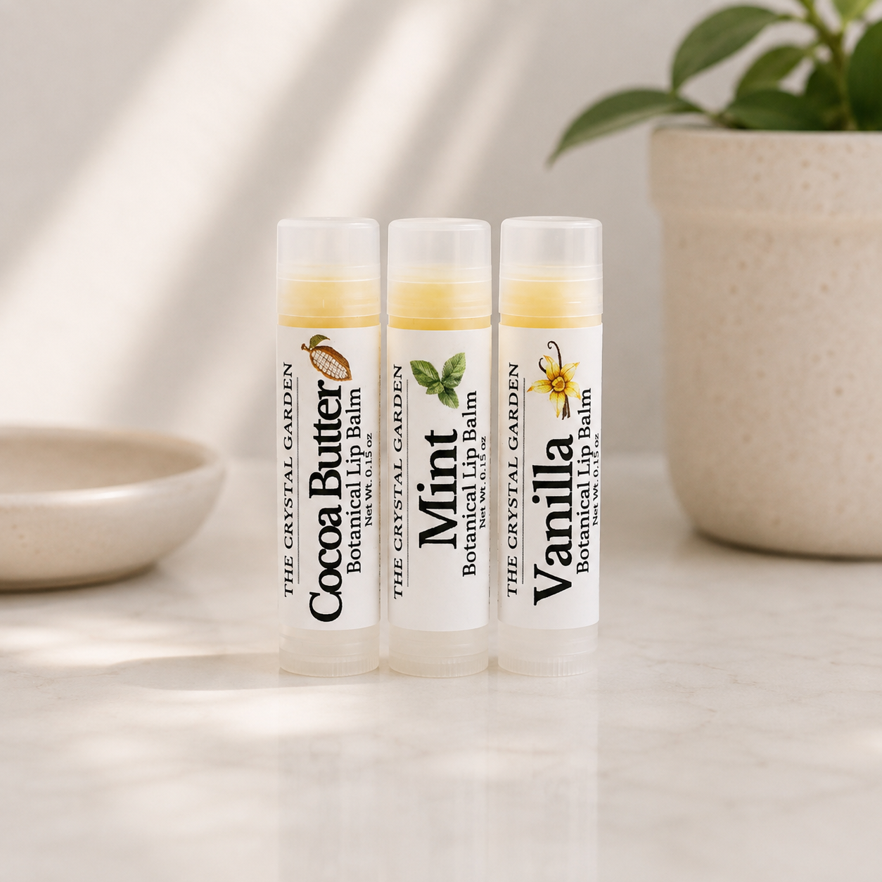 Three lip balm tubes standing upright on a light marble surface, with a ceramic bowl on the left and a potted plant in the background. The tubes are labeled Cocoa Butter, Mint, and Vanilla by The Crystal Garden Botanical Lip Balm.