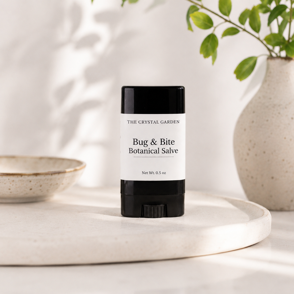 A black and white stick of 'The Crystal Garden' Bug & Bite Botanical Salve sits on a round white tray, styled with a ceramic bowl and a vase of green leaves in soft, natural light.