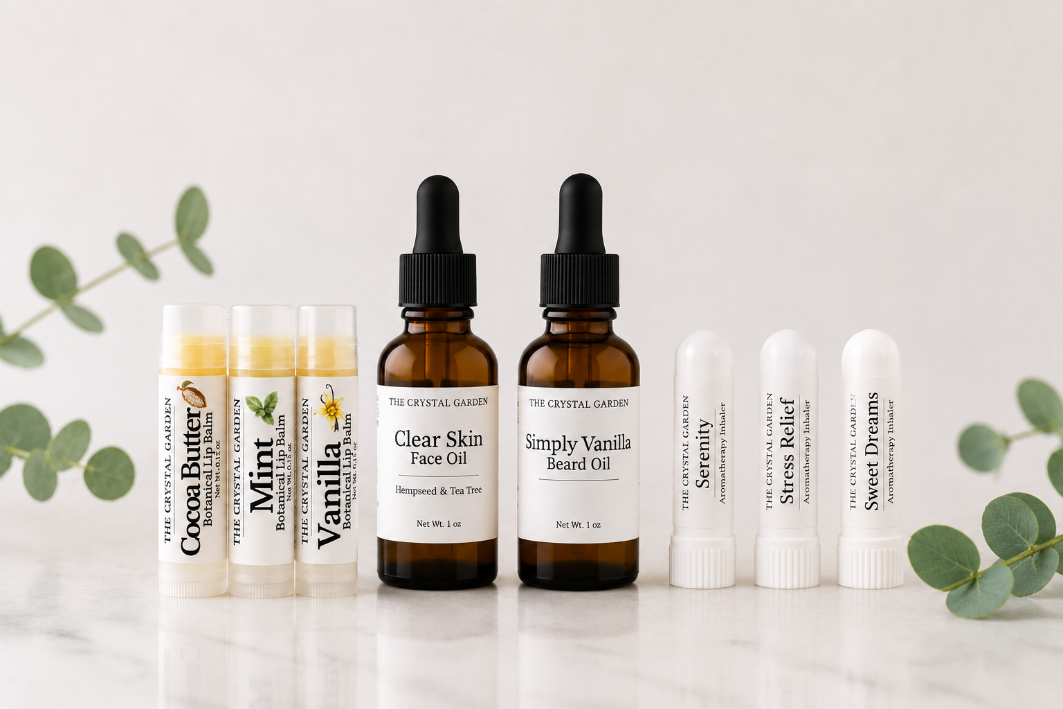 A set of facial and body care products from The Crystal Garden arranged on a light marble surface with eucalyptus leaves. The collection includes three amber dropper bottles (Clear Skin Face Oil, Simply Vanilla Beard Oil, and Unscented & Tea Tree), three white tubes (Cocoa Butter, Mint, and Vanilla), and three white lip balm tubes (Soothing, Stress Relief, and Sweet Dreams).