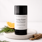A 1 oz stick deodorant from The Crystal Garden in the Cedar Forest Natural Deodorant scent, displayed on a round white ceramic tray with a slice of orange, greenery, and wood sticks against a white marble background.