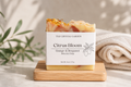 A rectangular bar of botanical soap from The Crystal Garden, labeled 'Citrus Bloom Orange & Bergamot', resting on a small wooden block. The soap has a white label and a top carved with orange and bergamot zest, set against a soft beige background with a folded towel and green leaves.