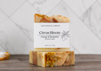 Two rectangular bars of The Crystal Garden Citrus Bloom botanical soap stacked on a wooden surface, with a white paper label on the top bar. The soap has a warm cream and light orange marbled body with dried orange flower petals embedded near the top and bottom ends.