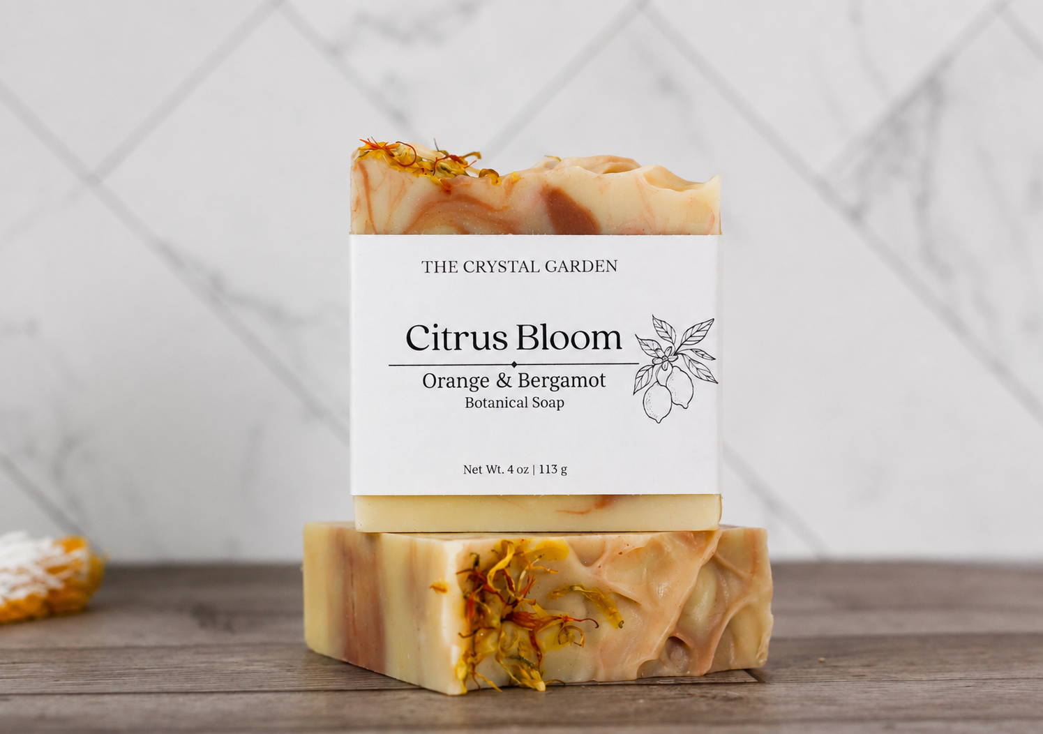Two rectangular bars of The Crystal Garden Citrus Bloom botanical soap stacked on a wooden surface, with a white paper label on the top bar. The soap has a warm cream and light orange marbled body with dried orange flower petals embedded near the top and bottom ends.