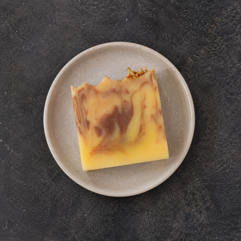 A square bar of marbled yellow and brown soap rests on a small speckled beige ceramic plate, set against a dark textured slate background.