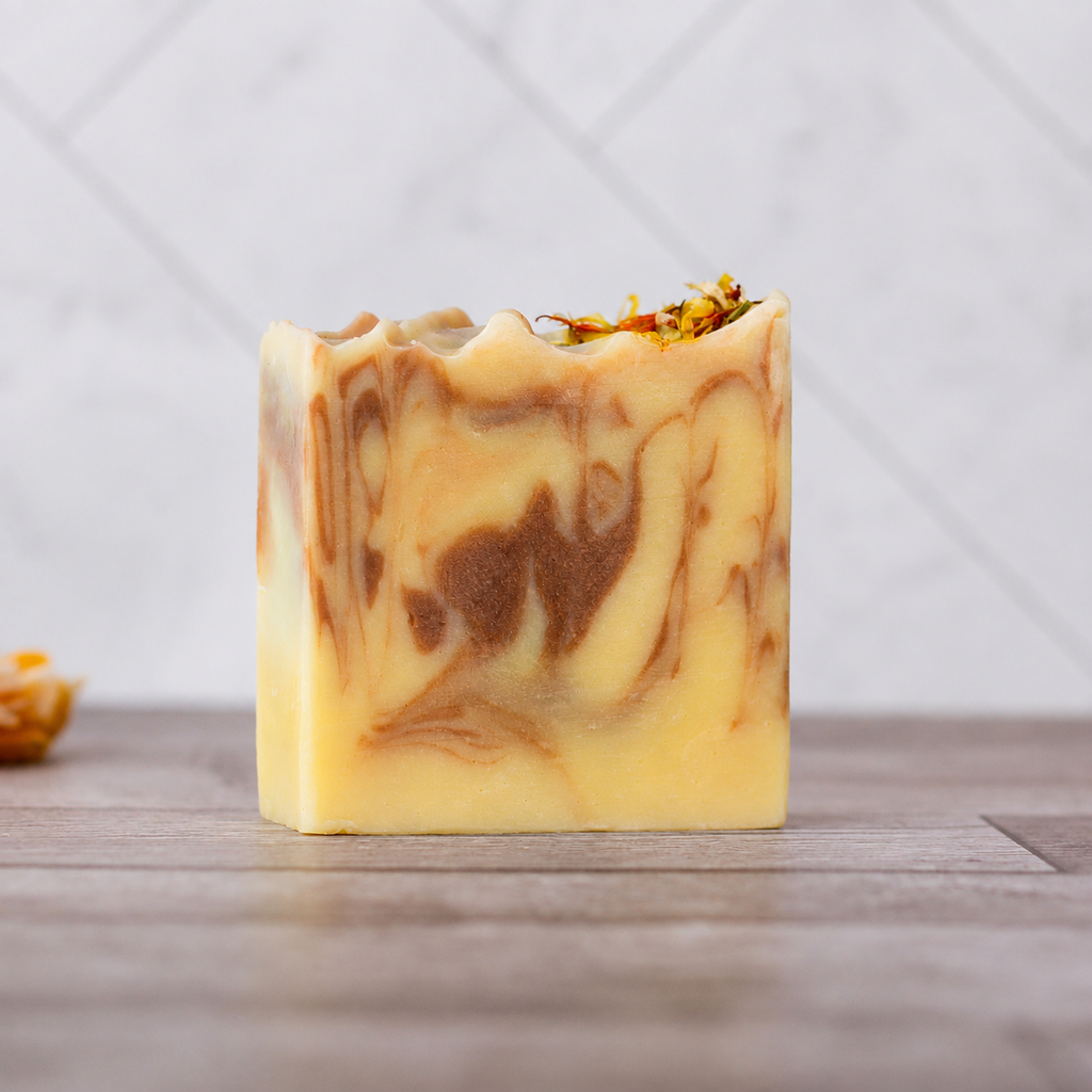 A single rectangular bar of handmade soap with a marbled swirl pattern of pale yellow, cream, and medium brown, topped with dried flower petals, standing on a wooden surface against a light gray tiled background.
