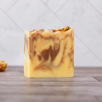 A single rectangular bar of handmade soap with a marbled swirl pattern of pale yellow, cream, and medium brown, topped with dried flower petals, standing on a wooden surface against a light gray tiled background.