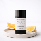 A black and white deodorant stick labeled 'Citrus Grove Natural Deodorant' by The Crystal Garden, styled on a light beige ceramic tray with orange and lemon slices against a white background.