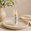 A lip balm tube labeled 'Citrus Mint' by The Crystal Garden, placed on a speckled beige ceramic plate with a neutral beige background, a vase with green leaves, and soft cream fabric.