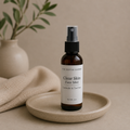 An amber glass face mist bottle with a white label and black spray top, standing on a small beige ceramic dish. A folded beige fabric and blurred green leaves in a vase appear in the background against a warm beige studio backdrop.