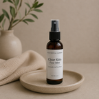 An amber glass face mist bottle with a white label and black spray top, standing on a small beige ceramic dish. A folded beige fabric and blurred green leaves in a vase appear in the background against a warm beige studio backdrop.