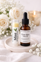 Two skincare products from The Crystal Garden, a Mint Botanical Lip Balm and Clear Skin Face Oil, displayed on a small white ceramic tray with white roses and a lit candle in the background.